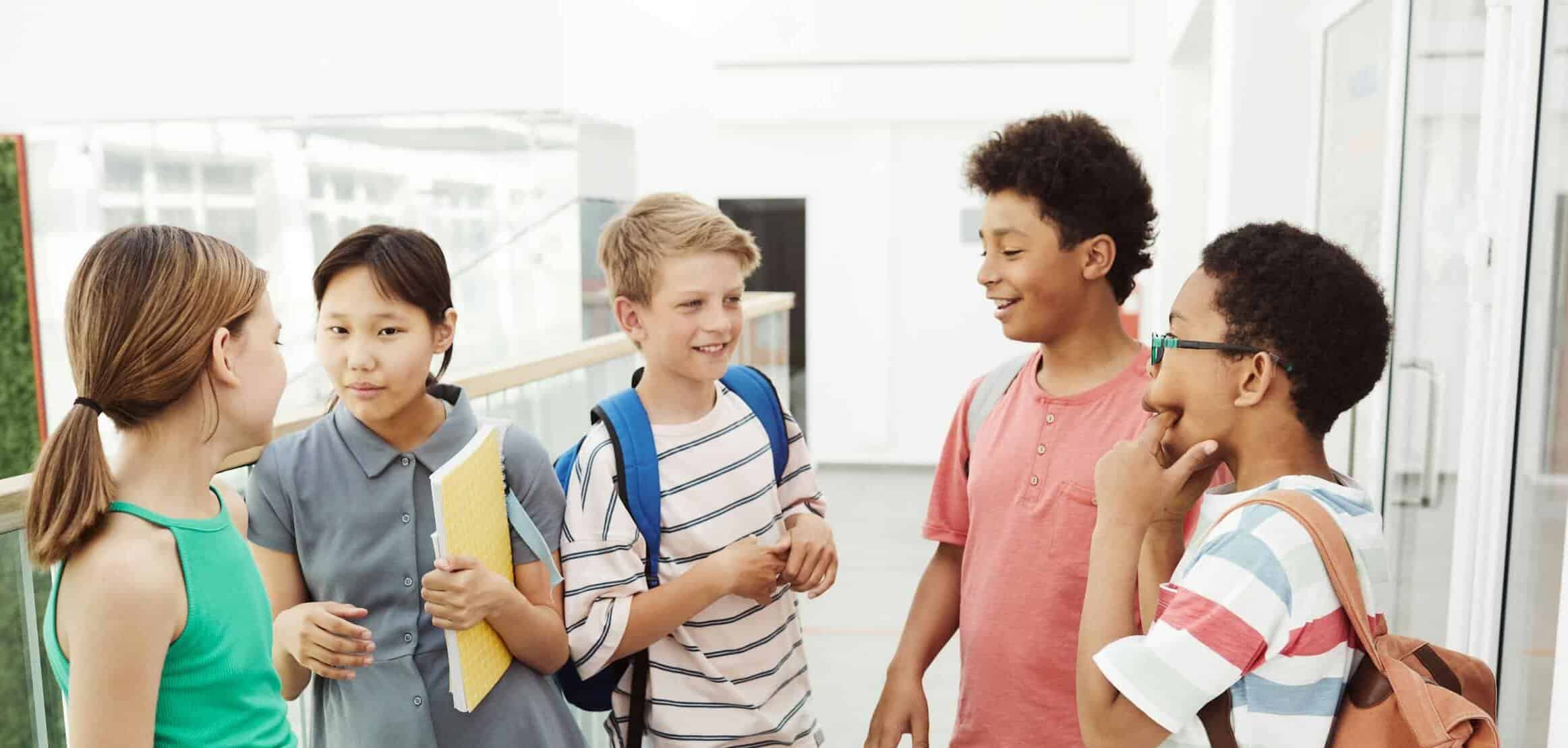 Diverse group of students talking and laughing in a school hallway, holding books and bags.